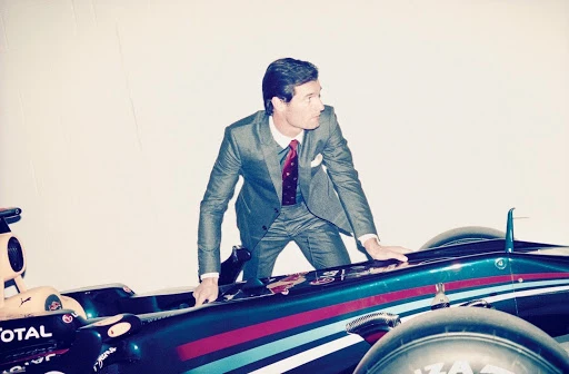 Man in a suit leans on a green and red Formula 1 race car in a photo studio, posing for a fashion editorial. 