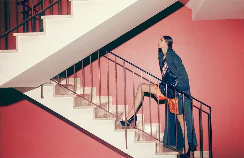 A woman in stylish clothing climbs a modern staircase with pink walls. She is wearing a coat and heels, ascending the white steps.