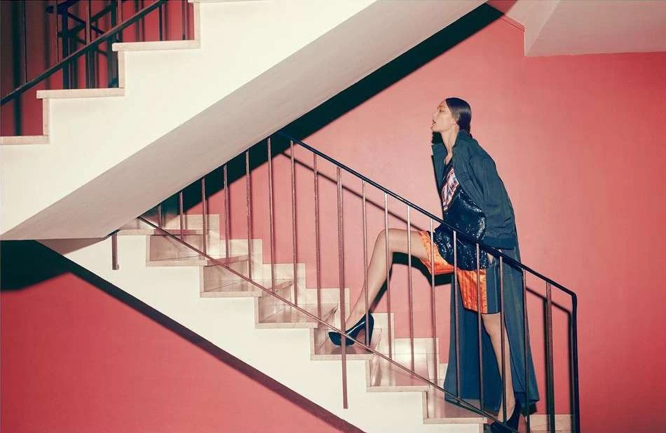 A woman in stylish clothing climbs a modern staircase with pink walls. She is wearing a coat and heels, ascending the white steps.
