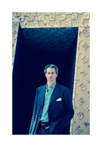 Portrait of an adult man in a blue blazer standing in front of a dark opening with a floral patterned border in an indoor setting.