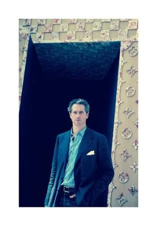 Portrait of an adult man in a blue blazer standing in front of a dark opening with a floral patterned border in an indoor setting.
