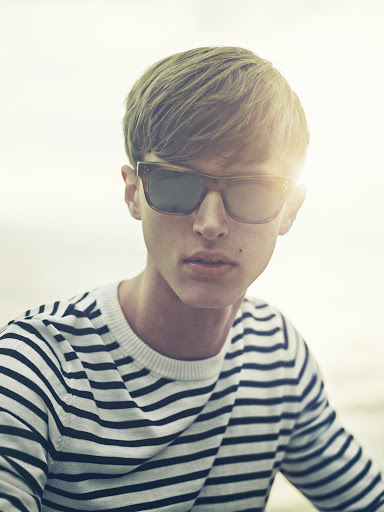 Portrait of a young adult male with blond hair and sunglasses wearing a striped sweater against a bright outdoor background.