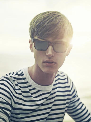 Portrait of a young adult male with blond hair and sunglasses wearing a striped sweater against a bright outdoor background.