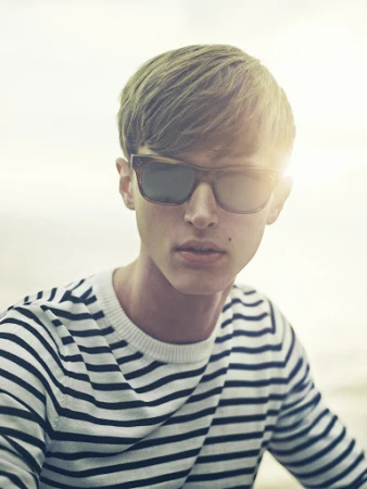 Portrait of a young adult male with blond hair and sunglasses wearing a striped sweater against a bright outdoor background.