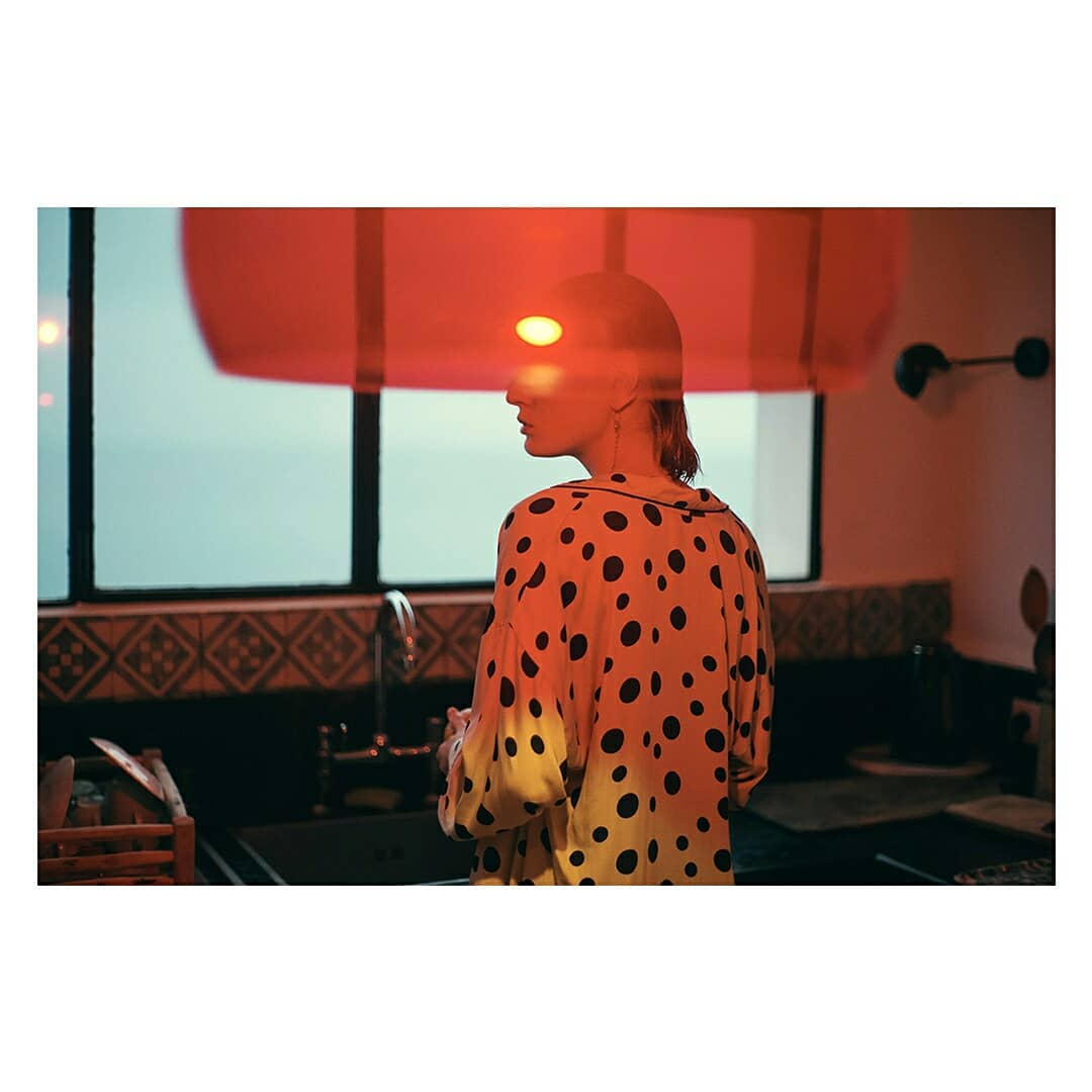 Woman in polka dot shirt stands near a kitchen sink and window, creating a double exposure effect with a red curtain and the outside.
