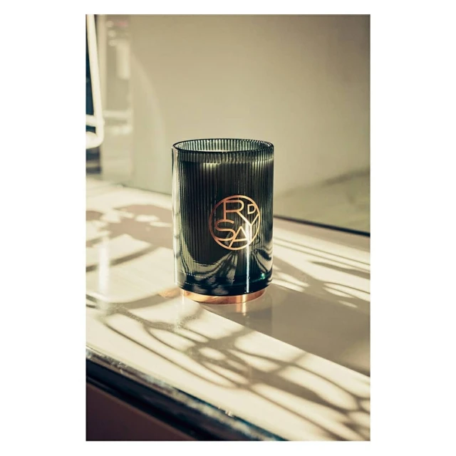 Dark green ribbed glass candle with a gold logo and base is sitting on a white surface with shadows, creating an elegant, minimalist scene.