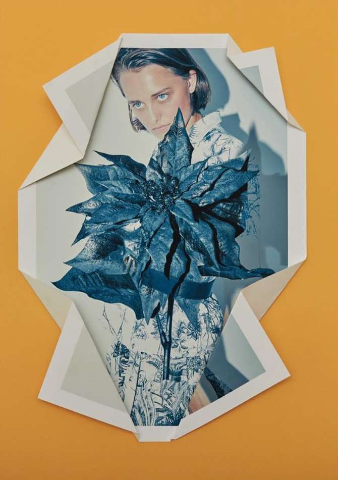 Artistic photo of a woman wearing a floral dress, framed by folded paper edges, with a large blue poinsettia flower in the foreground.