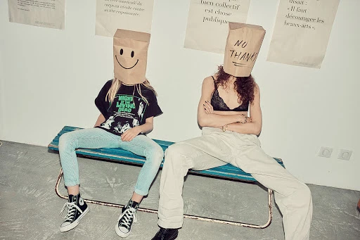 Two people wearing paper bags over their heads sit together on a blue bench against a plain white wall with posters