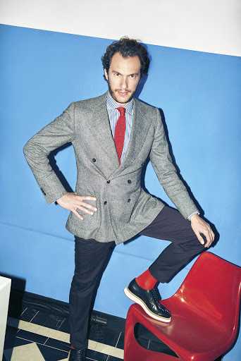Man in grey tweed jacket, blue striped shirt, and red tie poses with one foot on a red chair against a blue wall in an indoor studio.
