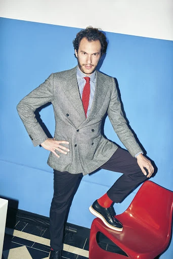 Man in grey tweed jacket, blue striped shirt, and red tie poses with one foot on a red chair against a blue wall in an indoor studio.