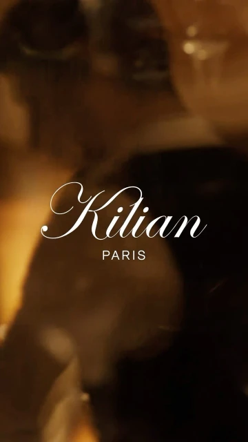 Close-up studio shot of Kilian Paris logo in script font on blurry brown and black background. Elegant beauty advertising.