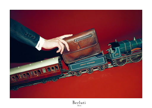 A brown leather Berluti pouch is held above a toy train on a red surface, with a man in a suit visible. Elegant product shot.