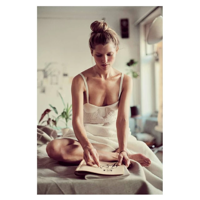 Woman with a top bun sits on a covered surface, holding a book, with a light and airy interior, creating a calm and relaxed scene.