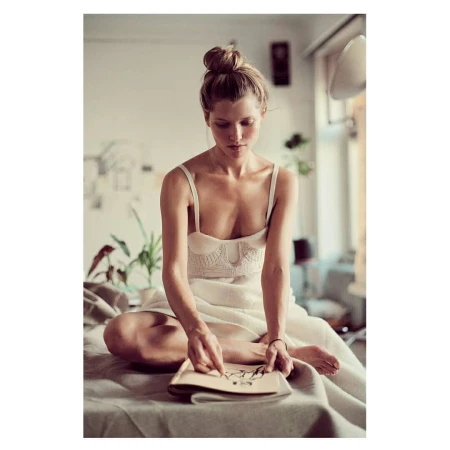 Woman with a top bun sits on a covered surface, holding a book, with a light and airy interior, creating a calm and relaxed scene.