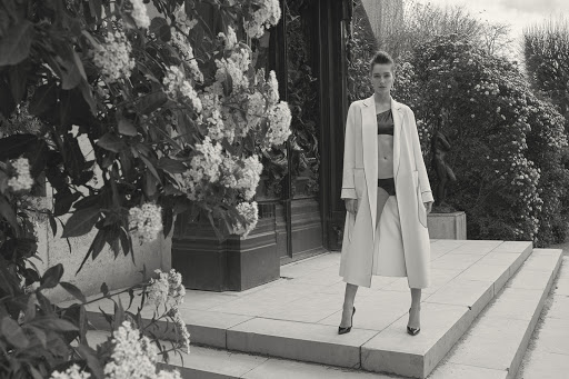 Black-and-white fashion shot of a woman in an open coat standing on stone steps with flowering bushes in the background.
