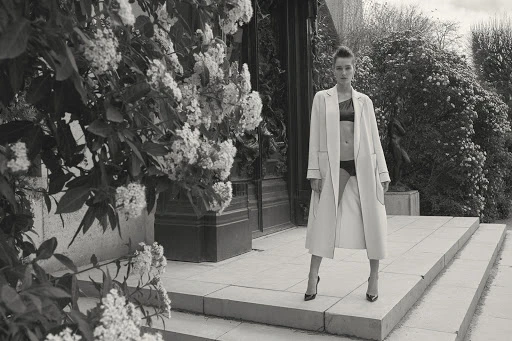 Black-and-white fashion shot of a woman in an open coat standing on stone steps with flowering bushes in the background.