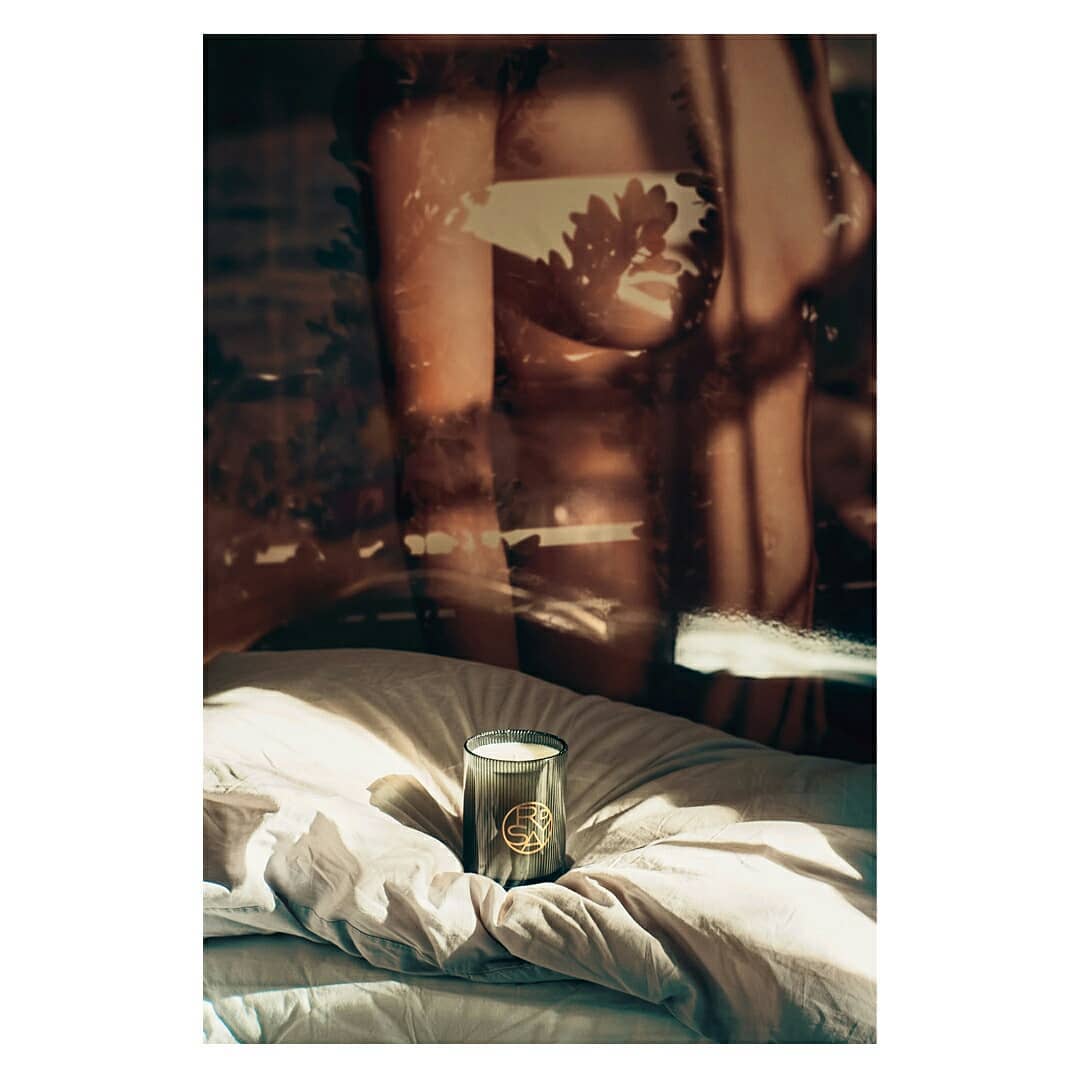 A lit candle sits on wrinkled bedsheets in soft sunlight, with a semi-nude figure visible in the background through a window or reflection.