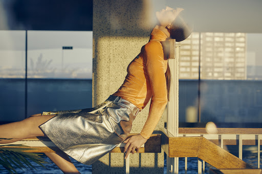 Woman in a metallic silver skirt and orange top poses against a backdrop of blue water and tall buildings in an urban setting.