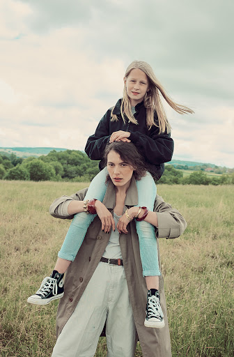 Woman with child on her shoulders stands in a grassy field with trees and cloudy sky in the background; they both wear sneakers.
