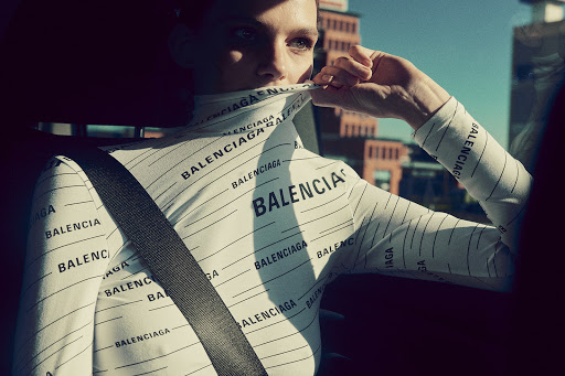 Indoor shot of a woman wearing a white Balenciaga turtleneck and a seatbelt against an out-of-focus urban backdrop, fashion-forward and chic.