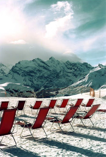 Red deck chairs on snow overlooking a snow-covered mountain range against a cloudy sky on a sunny day in a winter landscape.