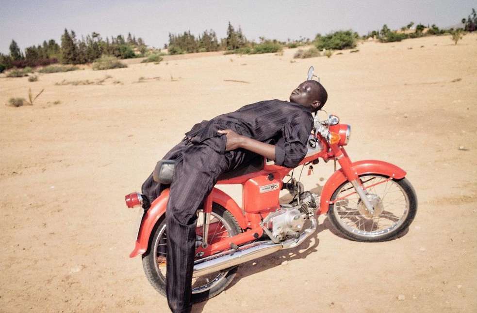 An adult male rests on a red vintage motorcycle outdoors in a desert environment during daytime, wearing a dark button-down shirt and pants.