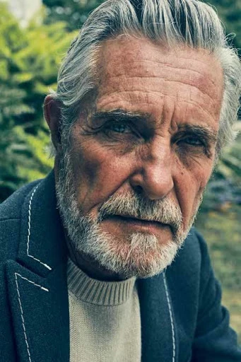 Portrait of an elderly man with gray hair and beard, wearing a blazer, gazing directly at the camera in an outdoor setting.