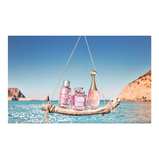Three perfume bottles displayed on a wooden plank suspended over a sparkling blue sea against a clear sky and coastal cliffs.