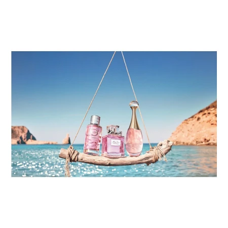 Three perfume bottles displayed on a wooden plank suspended over a sparkling blue sea against a clear sky and coastal cliffs.