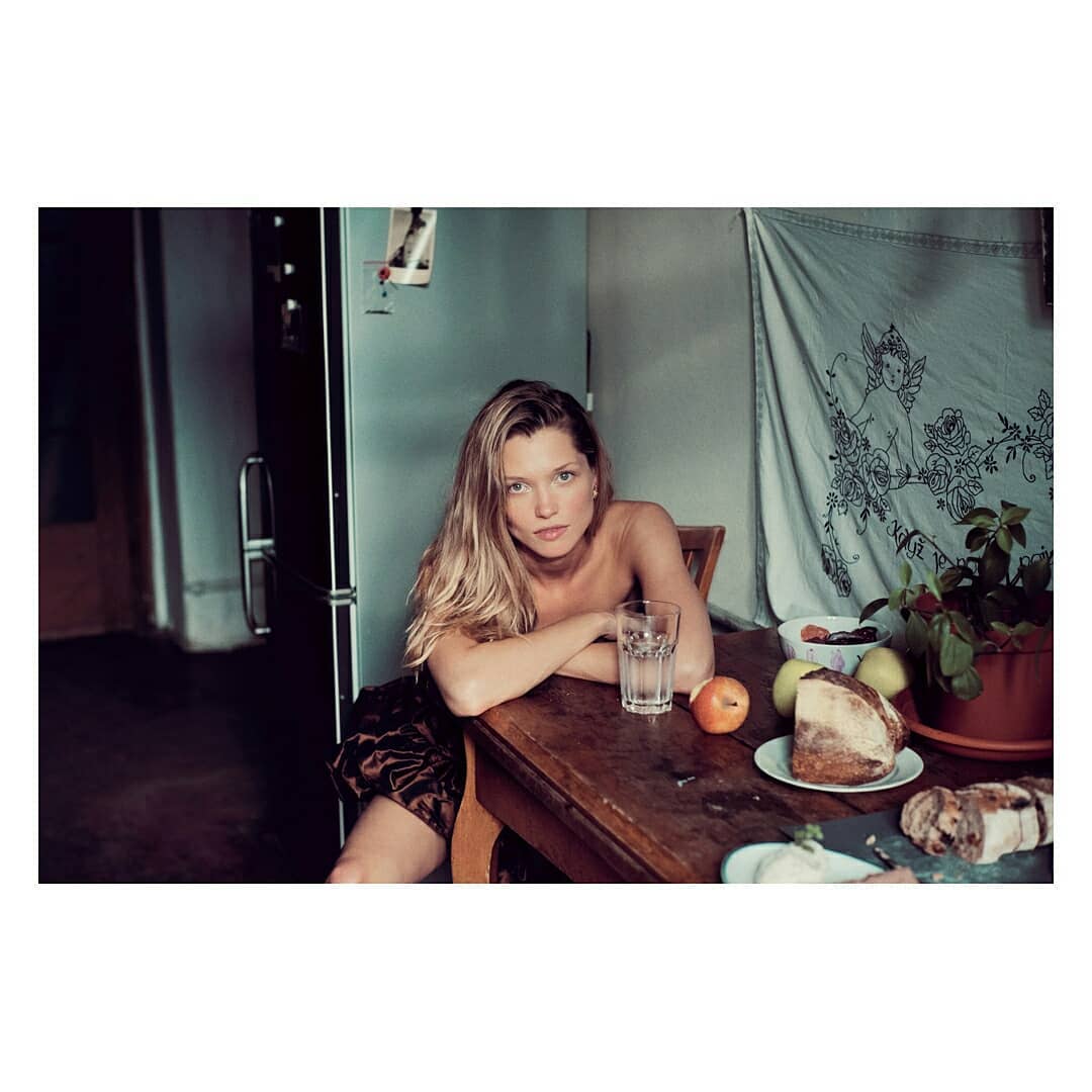 Woman leaning on a wooden kitchen table with a glass of water and bread loaves, photographed by Arnaud Pyvka.