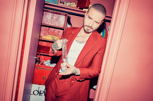 Adult male in red suit poses in closet holding a large chain and pendant. The closet contains clothes and boxes.