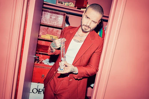 Adult male in red suit poses in closet holding a large chain and pendant. The closet contains clothes and boxes.