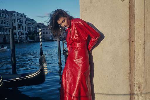 Woman in a red dress standing next to a stone wall in Venice, Italy, with a gondola and canal in the background.