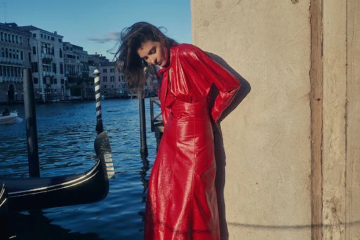 Woman in a red dress standing next to a stone wall in Venice, Italy, with a gondola and canal in the background.