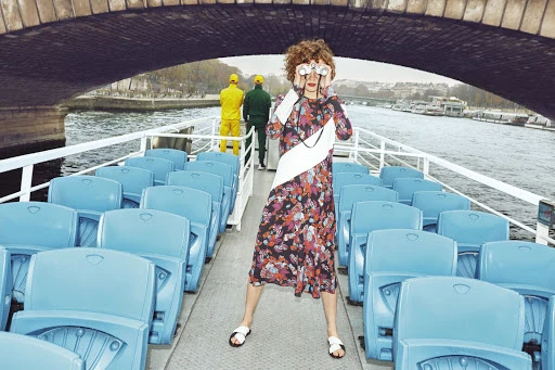 Woman in floral dress stands on a boat looking through binoculars on the Seine River in Paris with a bridge overhead.