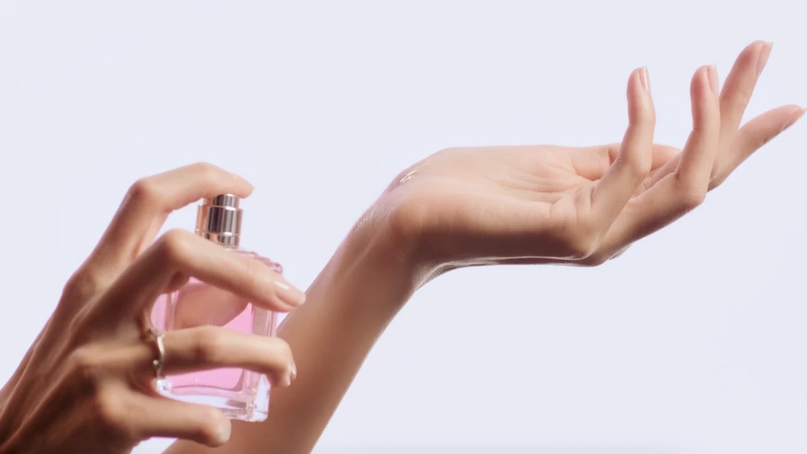 A woman's manicured hand sprays pink perfume from a glass bottle onto her other hand against a clean, bright white background.
