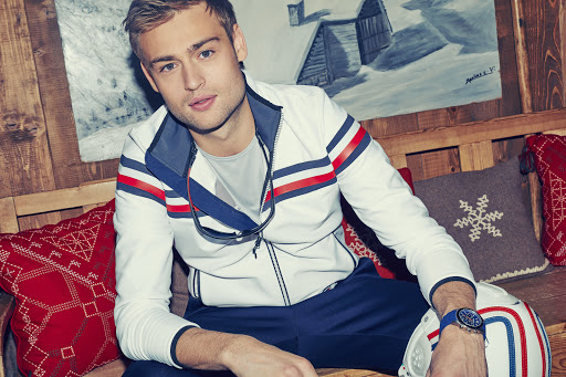 Portrait of a handsome man in a white athletic jacket, seated indoors with pillows, painting, and wood paneling in the background.