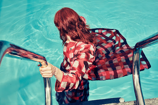 Red-haired woman in a checked red and white top exiting a blue swimming pool, holding onto metal handrails.