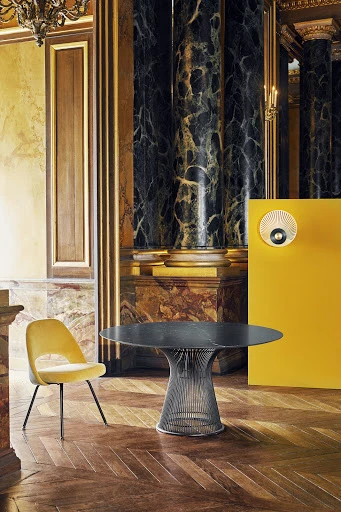 Interior shot of a round table with a metal frame and a chair with yellow fabric, set against a marble wall and parquet floor.