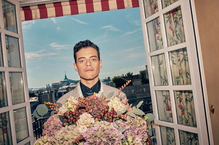 Portrait of Rami Malek in front of an open window overlooking Paris, holding a large bouquet of flowers, wearing a suit and black shirt.