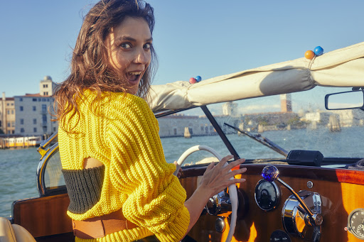 Woman with open mouth at boat steering wheel with Venice cityscape in background on sunny day; wearing yellow knit sweater. 