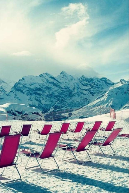 Many pink folding chairs stand in rows on snow with snow-covered mountains in the background under a blue sky with light clouds.