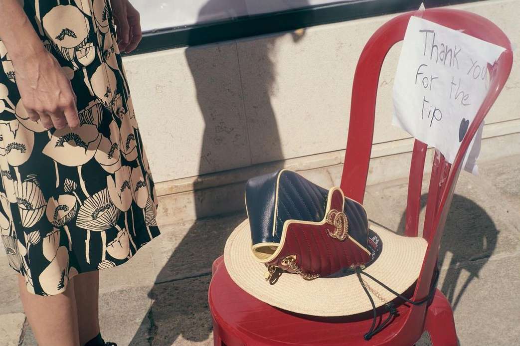 Handbag and straw hat rest on a red chair next to a woman in a floral-print dress, with a thank you note attached to the chair.