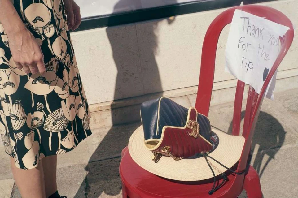 Handbag and straw hat rest on a red chair next to a woman in a floral-print dress, with a thank you note attached to the chair.