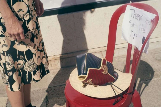 Handbag and straw hat rest on a red chair next to a woman in a floral-print dress, with a thank you note attached to the chair.