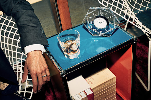 Close up of a man in a suit seated next to a blue side table with a glass of liquor, a crystal clock, and a stack of books.