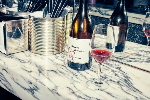 Wine bottles and wine glasses are placed on a marble countertop, alongside utensils in metal containers for a bar scene.