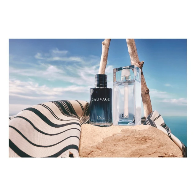 Two Dior fragrance bottles displayed on a stone surface with a striped cloth, set against a backdrop of a blue sky and ocean.