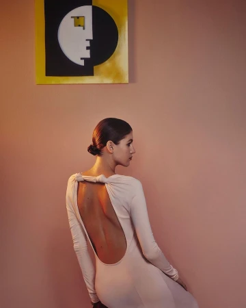 Back view of an adult woman in a light pink, open-back gown, posing in an indoor environment with modern art on the wall.