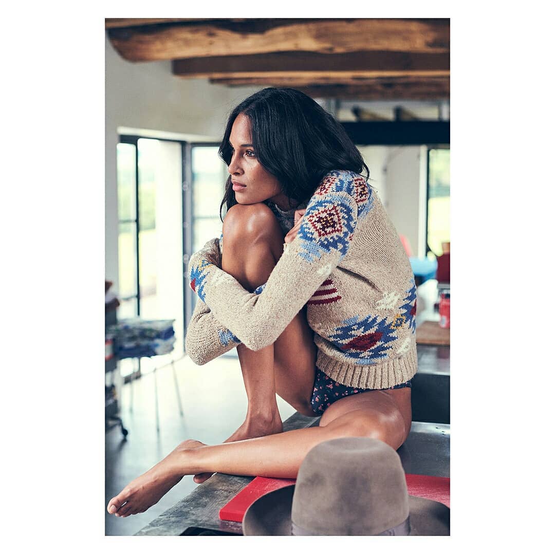 Adult female in a beige patterned sweater sitting on counter with knees up, looking off into the distance in an indoor environment.